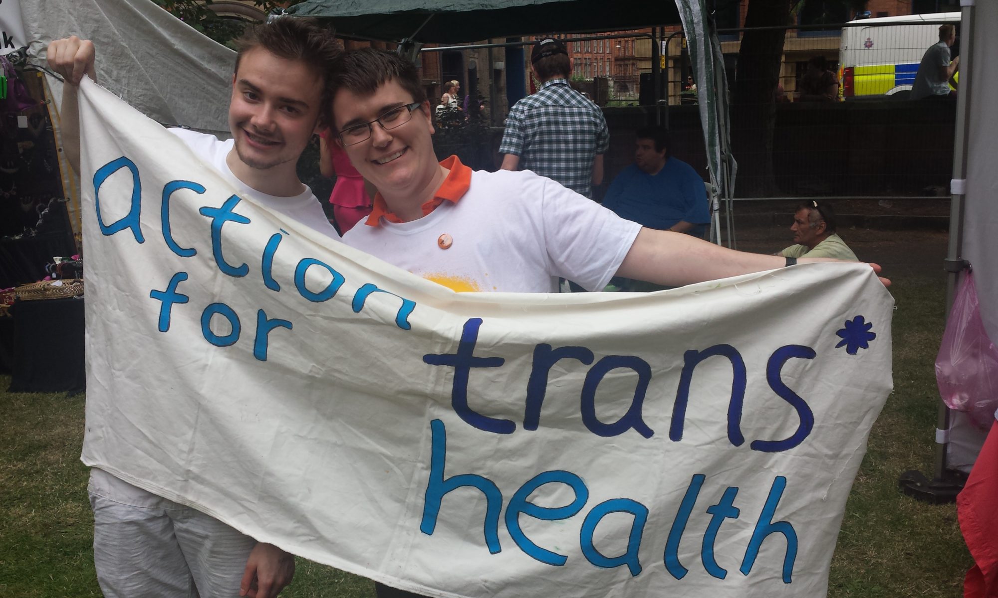 Two men in white t-shirts smiling while they hold an action for trans* health banner for the camera at an event in a park.